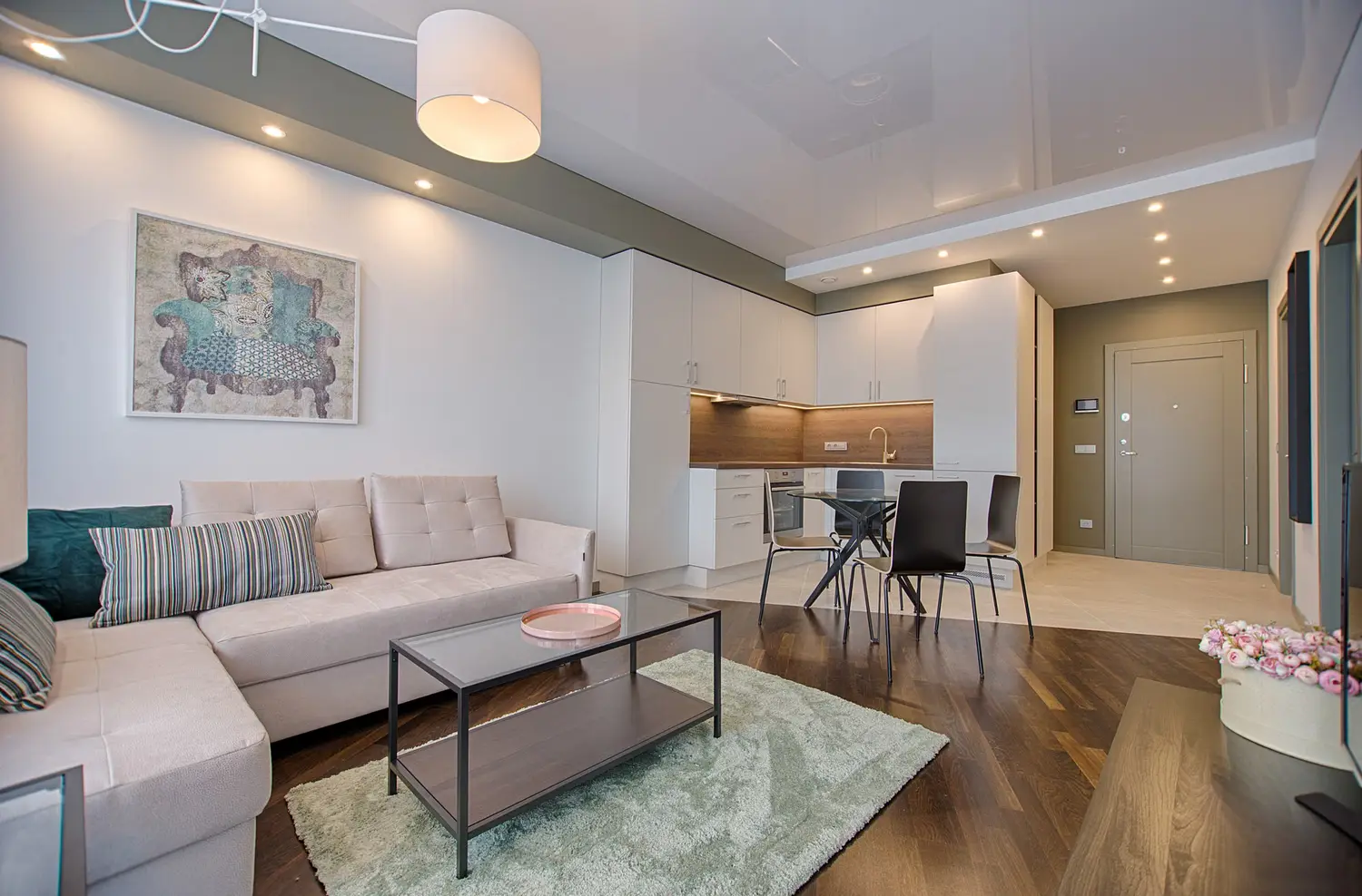 A compact apartment with a pale sofa, a glass coffee table, and a kitchen in the background