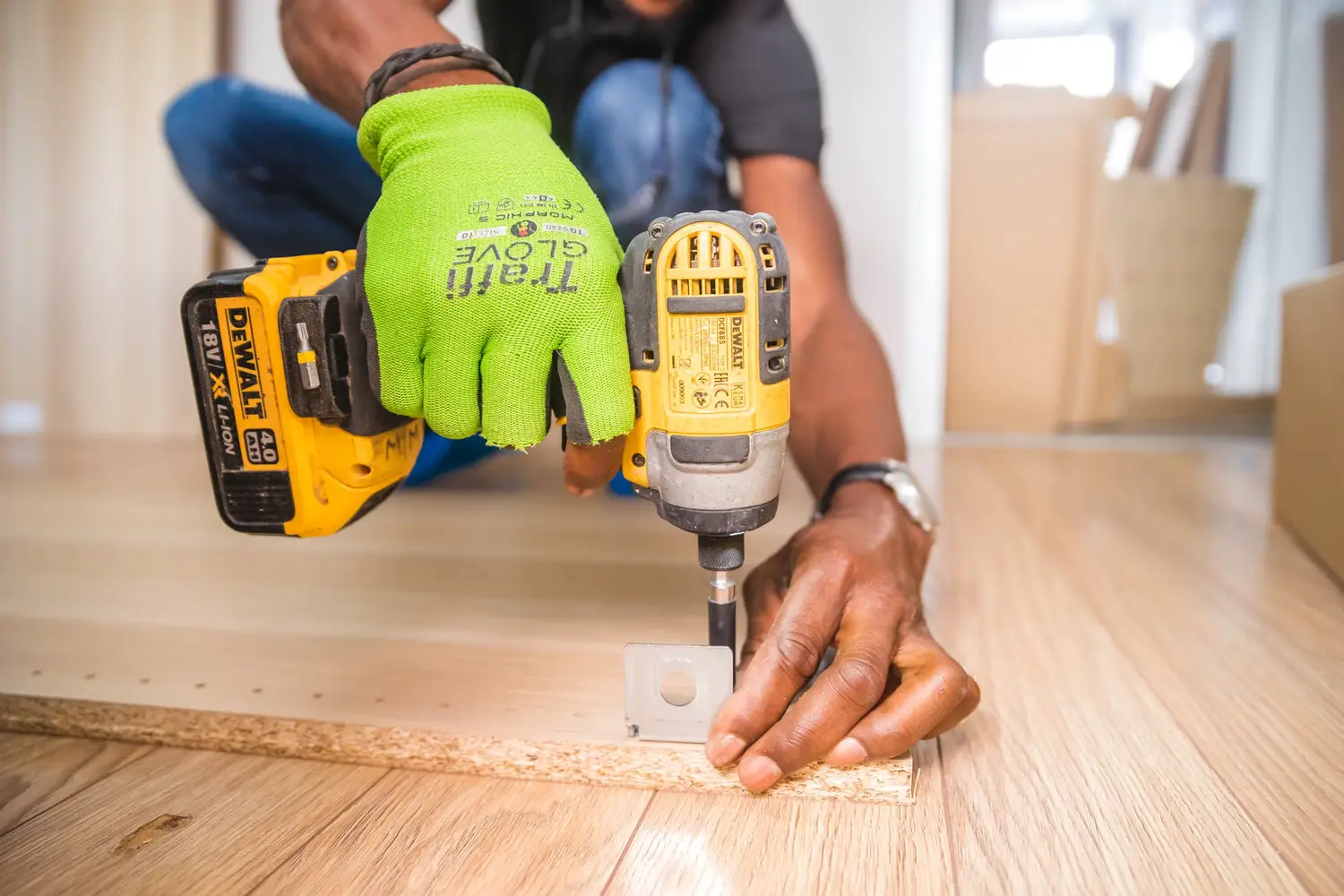 Close view of a gloved hand driving a screw into a particle-board panel laid on a wood floor