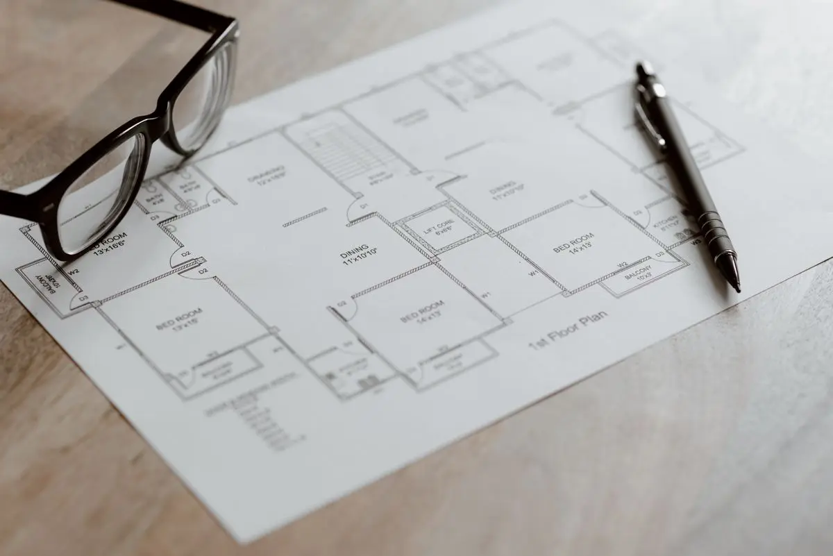 A printed one-story floor plan with room labels, glasses, and a pen on a wood surface