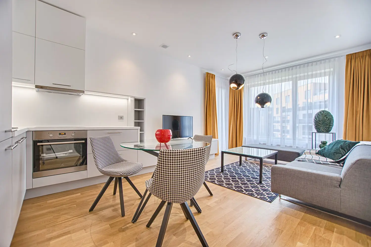 An open apartment with a glass dining table, four chairs, a gray sofa, and a kitchen along the left wall