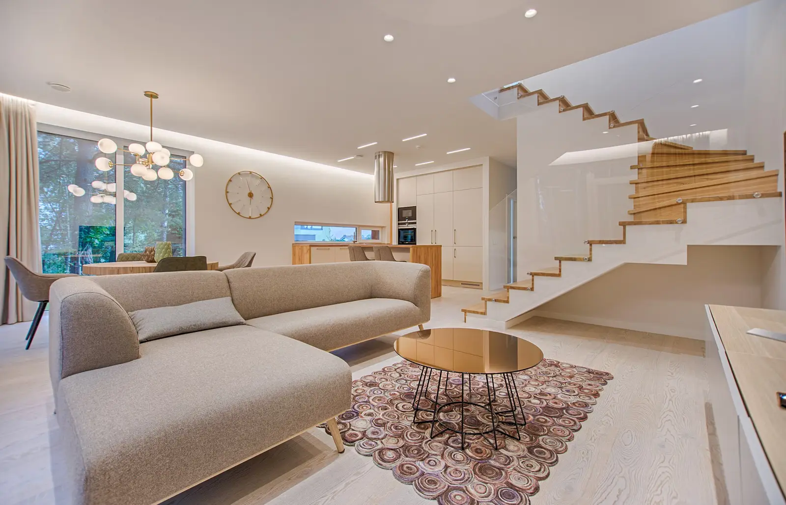 A light living room with a beige sectional sofa, a round metal coffee table, wood stairs, and built-in cabinets